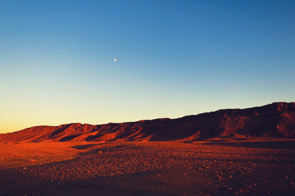 A photo of deep red cliffs across a sea of red sand. Above them, the sky is bright blue with a tiny moon visible.